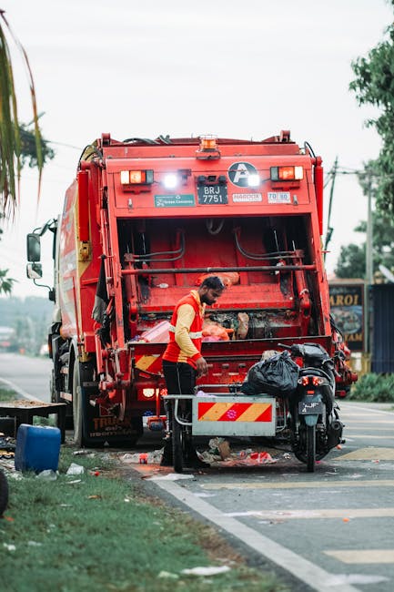 A waste collection worker wearing a yellow high-visibility vest and black trousers is engaged in rubbish removal, standing next to a large red waste collection vehicle parked on the side of a paved st