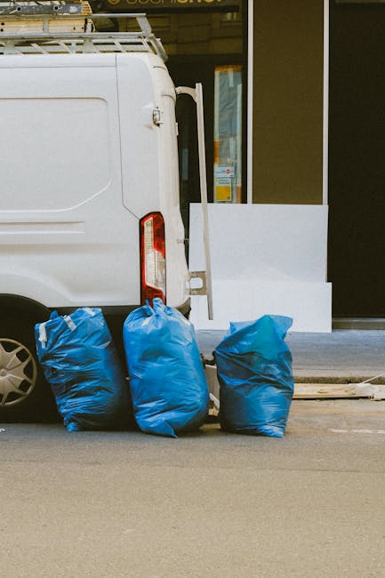 The image shows the rear section of a white commercial van parked on a street near a building with glass doors and windows. Adjacent to the van, on the pavement, are three large blue rubbish bags made