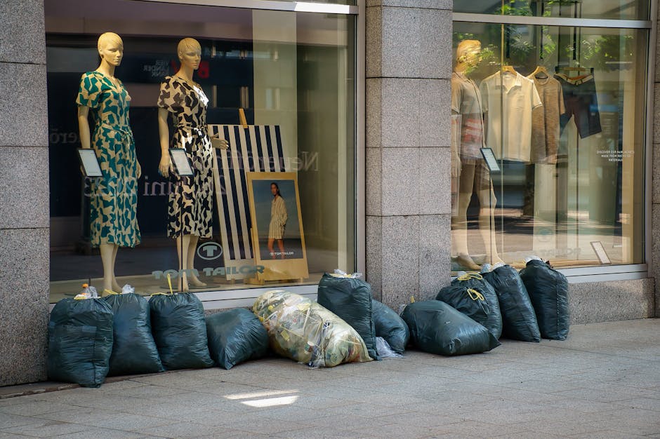 Three large black plastic sacks filled with waste material are placed on the pavement near a dark metal fence, which appears to be part of a residential or commercial property boundary. The sacks are 