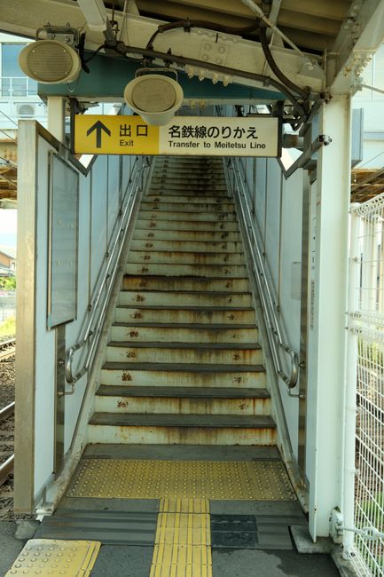A set of industrial stairs with black non-slip treads bordered by yellow safety strips, located indoors within a warehouse or factory environment. The stairs are supported by metallic side rails paint